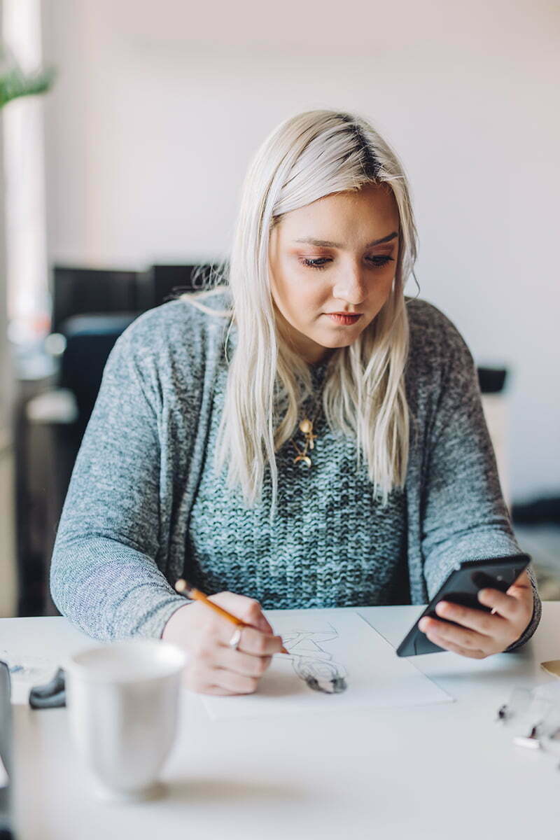Blond Woman Holding Mobile Phone and Pencil, Doing First Sketches On Paper At Table in Office
